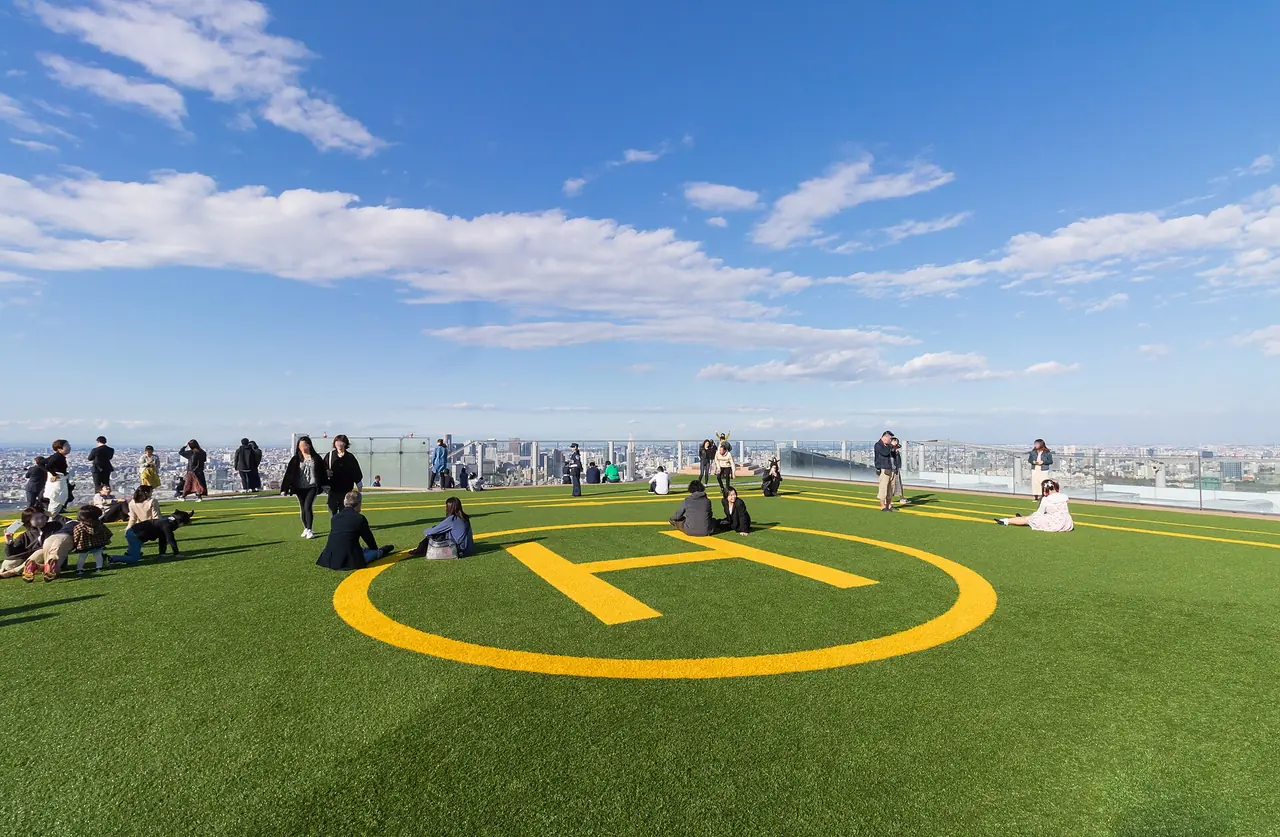 Visitors sitting and standing on the helipad lawn of the SHIBUYA SKY roof under a blue sky, with the yellow H marking