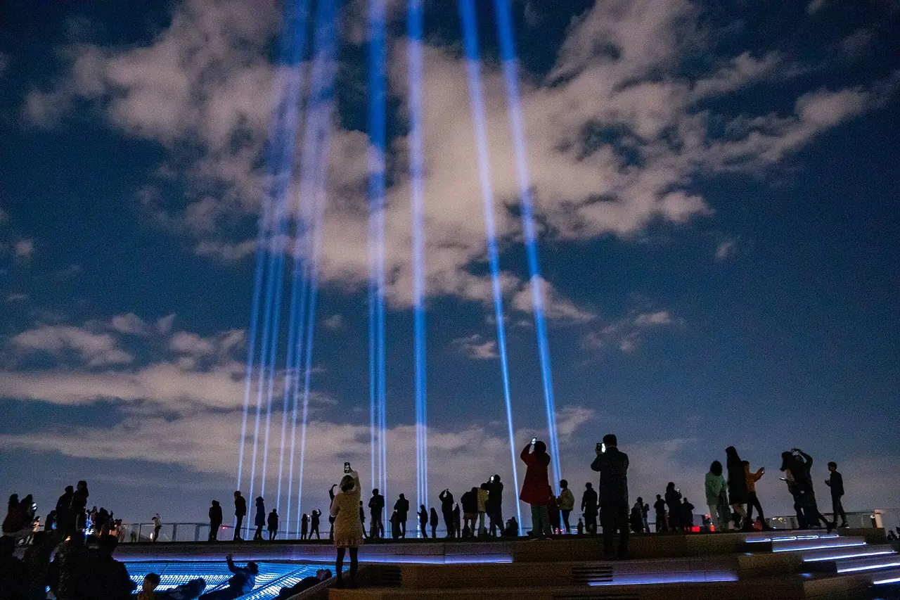 Blue Crossing Light beams rising from the SHIBUYA SKY roof into the evening sky, with visitors watching