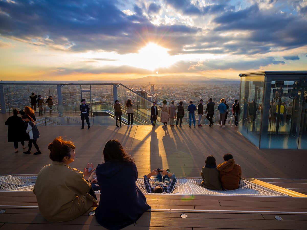 The open rooftop of SHIBUYA SKY at sunset, with people on the deck and the glass edge, and the sun low over the Tokyo skyline
